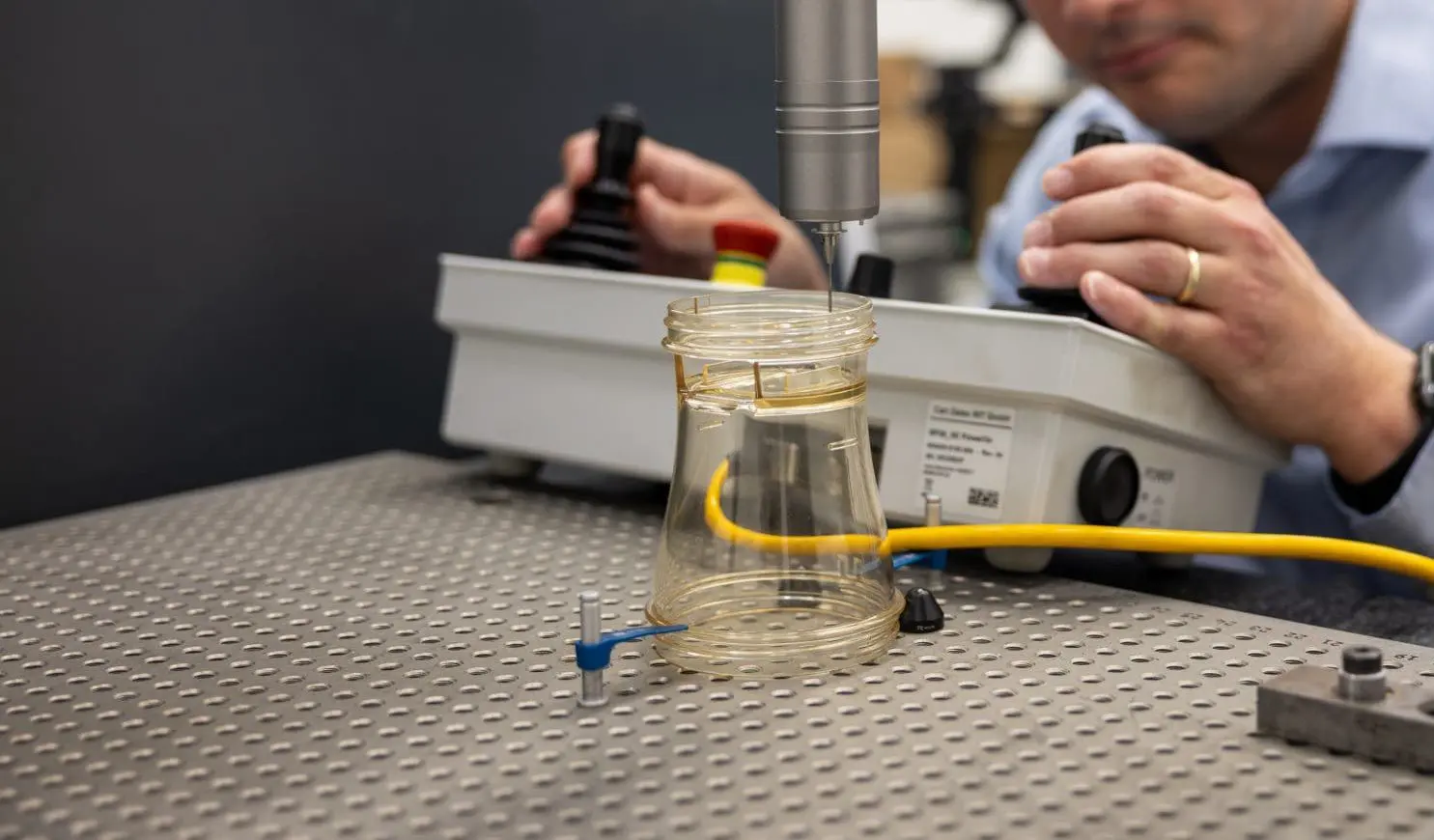 A person operates scientific equipment in a laboratory, focusing on a clear injection molded plastic container placed on a metal surface, with yellow and blue wires connected.