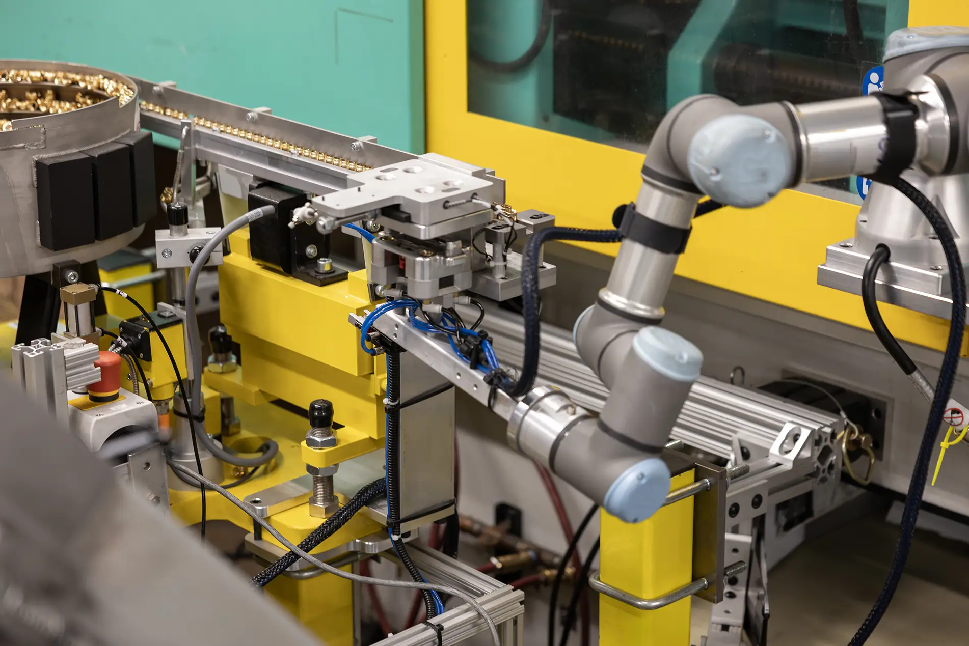 A robotic arm works on an automated assembly line in a factory, surrounded by yellow and metallic machinery parts, wires, and injection molded control equipment.