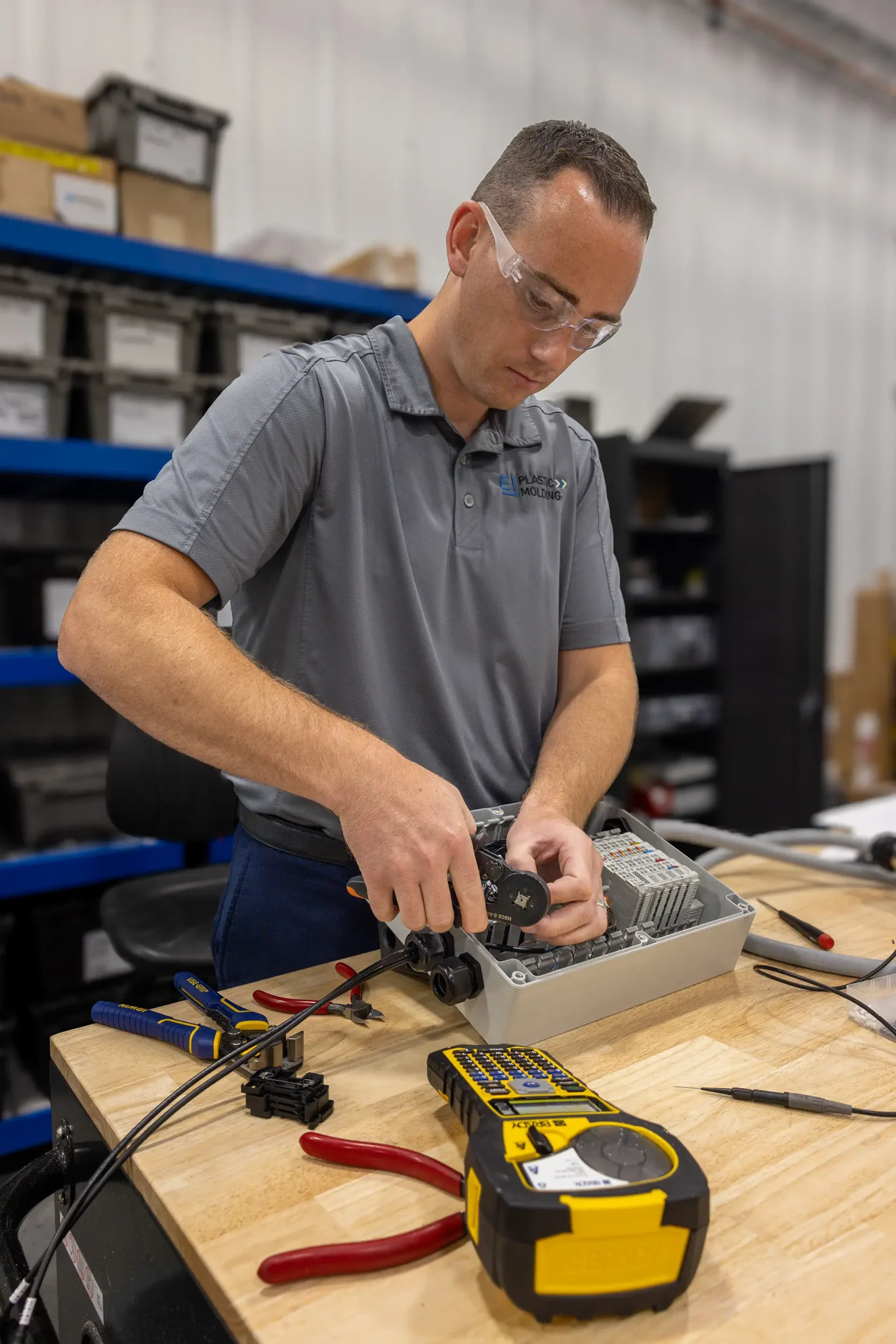 A man wearing safety glasses works on electronic equipment at a workbench, using a tool on a device. Various tools, including a digital multimeter and components related to plastic manufacturing, are on the table. Shelves and boxes are in the background.